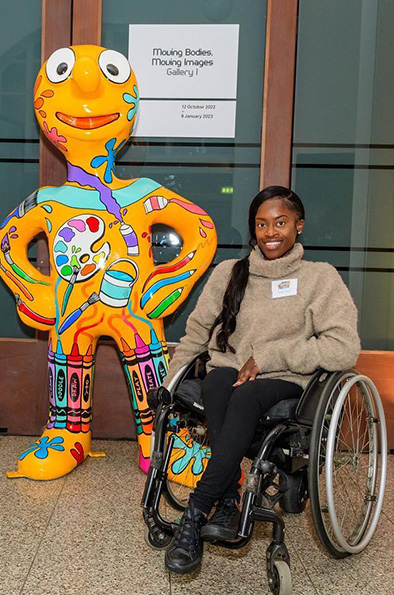 An orange painted Morph statue decorated with paint splashes and art equipment, a young female wheelchair user is posing next to him and smiling 